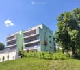 Modern multi-story apartment building with green and white exterior, multiple balconies, surrounded by trees and greenery.