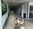 Balcony with tiled floor, scooter parked, surfboard leaning against scooter, covered area with concrete ceiling, glass railing, and outdoor furniture visible in the background.