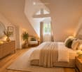 Cozy attic bedroom with slanted ceilings, large window, bed with pillows and blanket, nightstand with lamp, armchair, small table, mirror, and decorative plants.