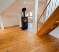 Bright hallway with wooden flooring, white walls, and a modern black wood-burning stove. A wooden staircase with a metal railing leads upwards.
