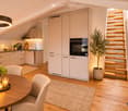 Modern kitchen and dining area with light-colored cabinets, a round wooden dining table, four chairs, a pendant light, and a staircase leading upwards.