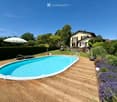 A wooden deck surrounds a swimming pool, with lounge chairs, an umbrella, and a house in the background.
