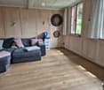 Cozy living room with wooden flooring, grey sectional sofa with pink and grey pillows, dartboard on wall, storage unit, and large windows with curtains.