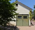 White stone building with a green garage door, surrounded by greenery and a paved pathway.