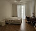 Bedroom with wooden floor, large window with white curtains, bed with striped blanket, desk with chair, and lamp.