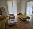 Living room with wooden flooring, large windows with white curtains, a sofa, a chair, a coffee table with a chessboard, and a small table with chairs.