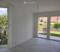 Empty room with white walls, tiled floor, large windows, and sliding glass doors leading to a patio and garden.