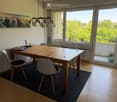 Dining room with a wooden table, chairs, a painting on the wall, and a hanging lamp. Large windows offer a view of greenery outside.