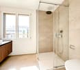 Modern bathroom with a glass-enclosed shower, a toilet, a sink with a wooden cabinet, and a large window offering a view of the city.