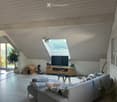 Modern living room with grey sofa, coffee table, TV, guitar, dog house, and large windows offering a scenic view.