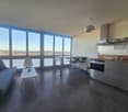 Modern open-plan living and dining area with a sofa, small table, dining table with chairs, and a kitchen area with an oven and cabinets. Large windows offer a scenic view.