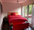 Cozy bedroom with a wooden bed frame, red bedding, a nightstand, and a red cabinet. Large windows offer a view of greenery outside.