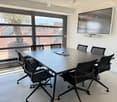 Conference room with a large table, multiple chairs, and a wall-mounted screen. Large windows offer a view of the surrounding area.