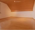 Empty room with wooden flooring and white walls, featuring a slanted wooden ceiling and a window with multiple panes.