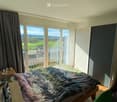 Bright bedroom with large windows offering a scenic view, featuring a bed with a colorful blanket, a nightstand, a wardrobe, a clothing rack, and a chair.