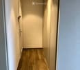 A narrow hallway with light wooden flooring, white walls, and recessed ceiling lights. The hallway features a closed door on the left and a mirrored wall on the right.