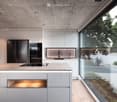 Modern kitchen with white cabinets, a large island, and a sleek black refrigerator. The space features a concrete ceiling with recessed lighting and expansive windows offering a view of the garden.