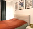 A modern bedroom with a neatly made bed, framed artwork, a nightstand with a decorative vase, and a floor lamp near the window.