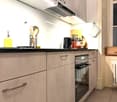 Modern kitchen with light-colored cabinets, stainless steel handles, and a built-in oven. Countertop features a bottle of olive oil, dish soap, and a basket of kitchen utensils. A wooden cutting board with knives and a black kettle are also visible.