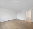 Empty room with light wooden flooring and white walls, featuring a closed door and an open doorway leading to a bathroom.