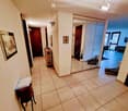 Hallway with tiled floor, white walls, and several doors leading to different rooms. A small table with papers and a framed map is visible on the left, and a mirror is mounted on the wall near the end of the hallway.