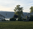 Lush green lawn with trees, bushes, and a trampoline, with houses and a distant mountain range in the background.