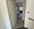 A modern hallway with tiled flooring, white walls, and a staircase leading to an open room with wooden flooring and large windows.