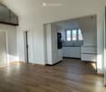 Bright, unfurnished room with wooden flooring, white walls, and large windows. Features a modern kitchen area with white cabinets and stainless steel appliances visible in the background.