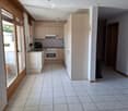 Bright kitchen with white tiled floor, light wooden cabinets, and stainless steel appliances including an oven and fridge. A large window with wooden frame provides natural light.