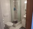 Modern bathroom with white tiled walls and floor, featuring a walk-in shower with glass enclosure, toilet, sink with mirror, and small cabinet.