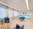 Modern, well-lit room with light wooden flooring, white walls, and large windows covered with sheer curtains. Features include a massage table, two black leather chairs, small wooden tables, a potted plant, a tall floor lamp, and a white cabinet with a co