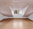 Empty room with wooden flooring and white walls, featuring a slanted wooden ceiling and large windows offering a view of the outside.