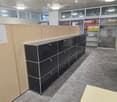 Modern office space with grey carpet, glass partitions, black metal cabinets, and shelves holding binders.