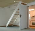 Open room with open staircase to upper floor, white walls, white door, gray flooring, ceiling light, kitchen in the background