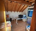 A furnished room with wooden beams on the ceiling, a desk with a laptop and screen, a chair, a small table with a cup, and a shelf with various items.