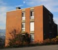 Modern two-story brick building with large windows and a flat roof, surrounded by greenery and shrubs.