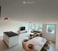 Modern living and dining area with a sofa, coffee table, dining table with chairs, and a kitchen island. Large windows offer a scenic view.