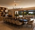 Elegant dining room with a long dark wooden dining table surrounded by beige upholstered chairs, a large chandelier, and decorative items on shelves. The room features a view of an outdoor dining area through large glass doors.