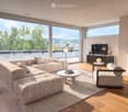 Modern living room with large windows offering a scenic view, a beige sectional sofa, a coffee table, an armchair, a television, a floor lamp, and a fireplace.
