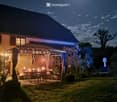 Nighttime view of a well-lit outdoor patio area with string lights, a dining table, and chairs, surrounded by greenery and a house in the background.