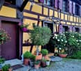 Traditional half-timbered house with yellow and purple exterior, surrounded by potted plants and shrubs in a gravel area.