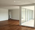 Empty room with wooden flooring, white walls, large glass sliding doors, and a visible adjacent room through an open doorway.