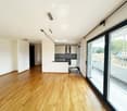 Bright, unfurnished room with wooden flooring, large glass sliding doors leading to a balcony, and a modern kitchen area visible in the background.