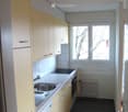 Modern kitchen with light-colored cabinets, stainless steel sink, oven, and radiator. Dark tiled floor and large windows.