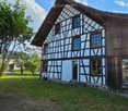 Traditional half-timbered house with light blue walls and dark wooden beams, surrounded by a grassy yard and trees.