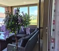 Balcony with a wicker sofa, glass table, and decorative plant with purple flowers, overlooking a scenic countryside view.