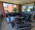 Balcony with a wicker sofa, glass table, two chairs, and flower pots, overlooking a building.