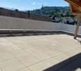 A spacious, sunlit balcony with a tiled floor and wooden beams, overlooking a scenic town with visible rooftops and distant mountains.
