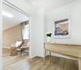 A modern, minimalist hallway with light wooden flooring, white walls, and a sleek console table. A framed poster is displayed on the wall, and a small vase with white flowers sits on the table. A wicker chair and small round table are visible in the adjac