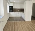 Empty kitchen with white cabinets, dark brown tiles, and wooden flooring. No appliances or furniture are present.